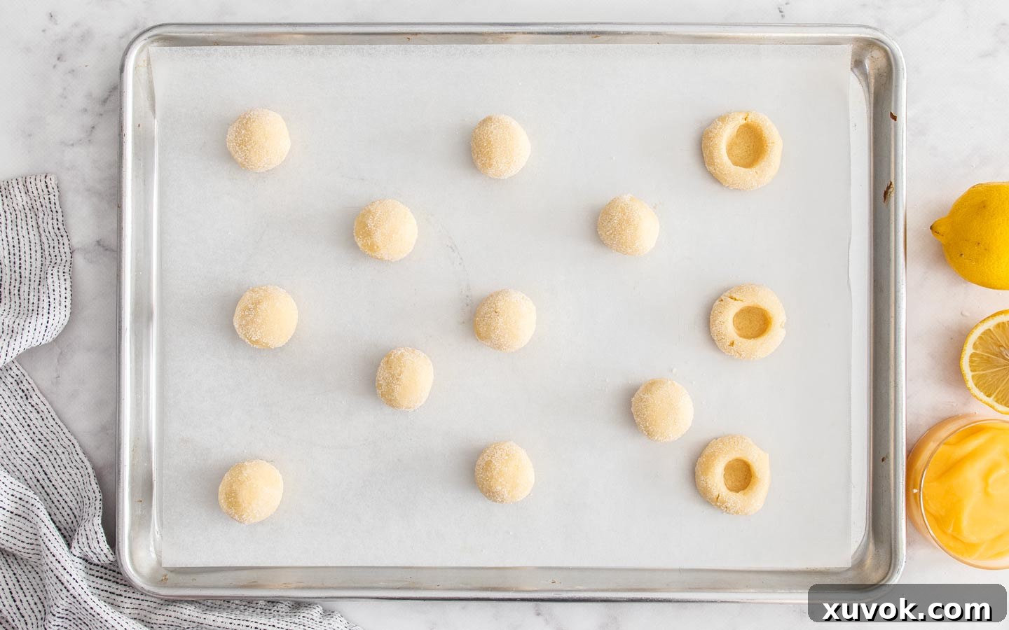 Several perfectly formed cookie dough balls on a baking tray, with some already featuring deep thumbprint indents, patiently awaiting their zesty lemon curd filling.