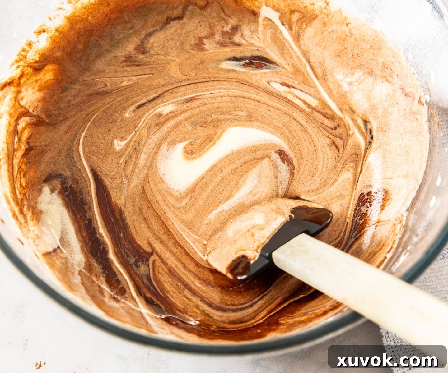 A spatula gently stirring the melted chocolate into the wet ingredients in a mixing bowl.