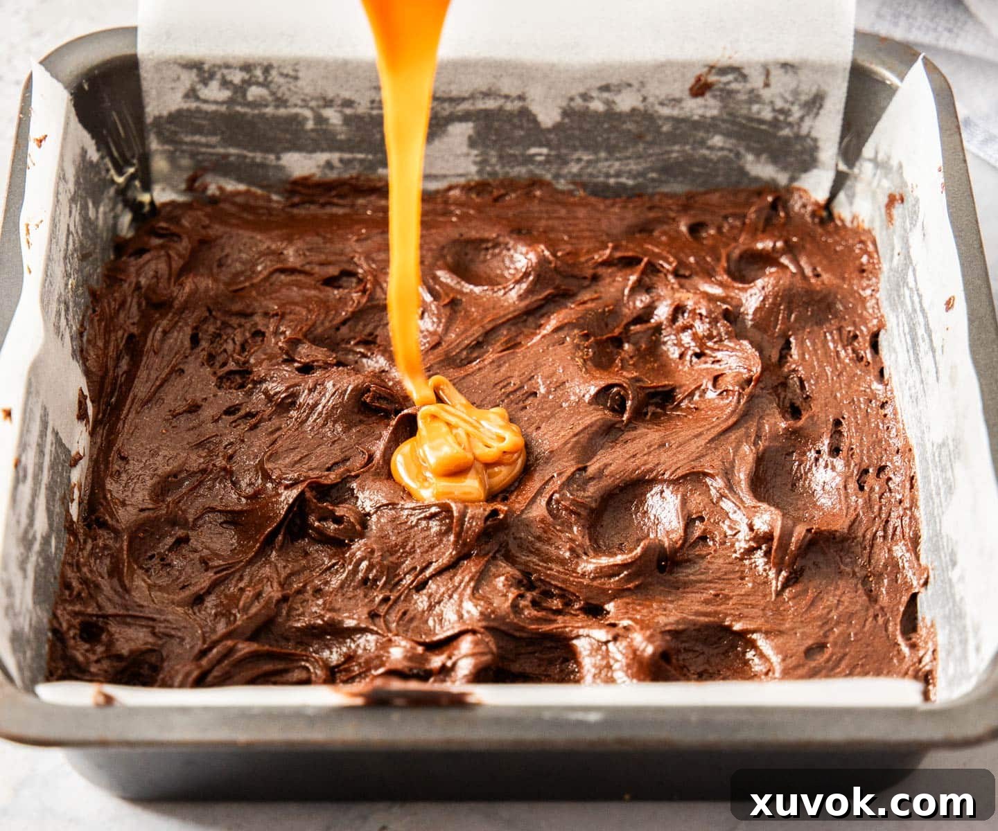 Golden caramel being poured and drizzled onto the first layer of chocolate brownie batter in a baking pan.