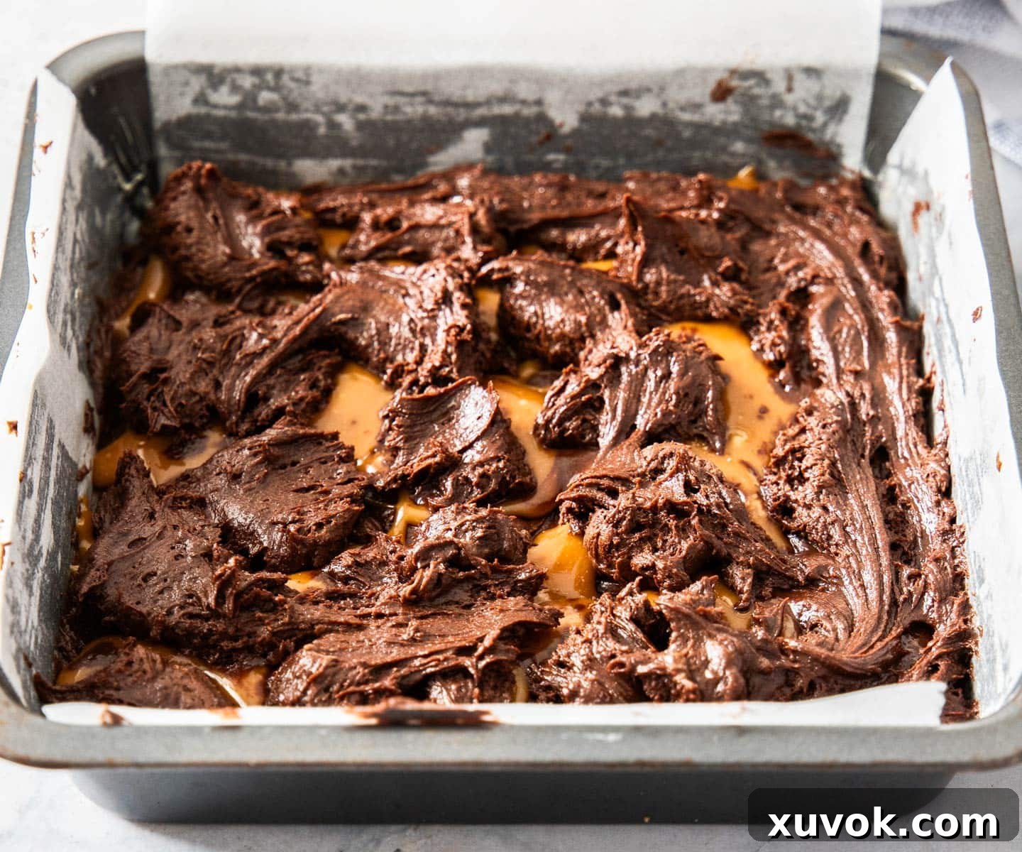 Brownie batter dolloped over the top of the caramel layer in a baking pan, creating a marbled effect.
