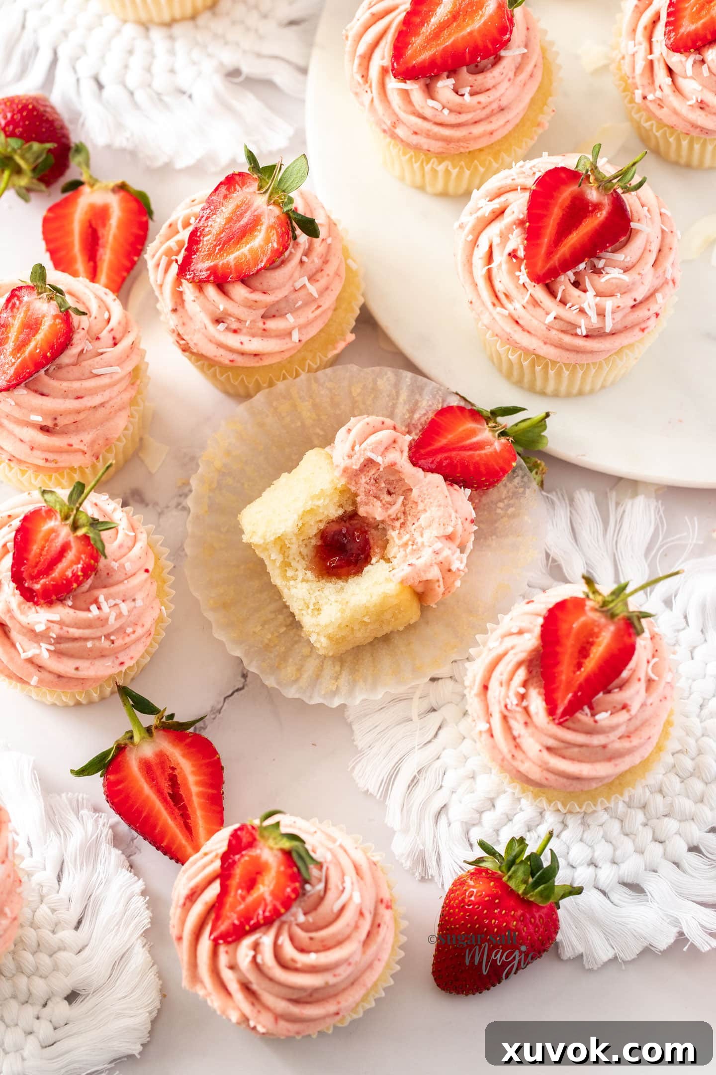 overhead view of a batch of cupcakes with strawberry jam in the middle, topped with pink frosting and a fresh strawberry