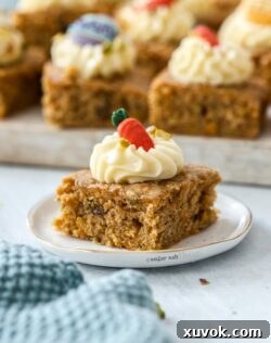 A closeup of a carrot cake bar on a small white plate. A tray of more in the background