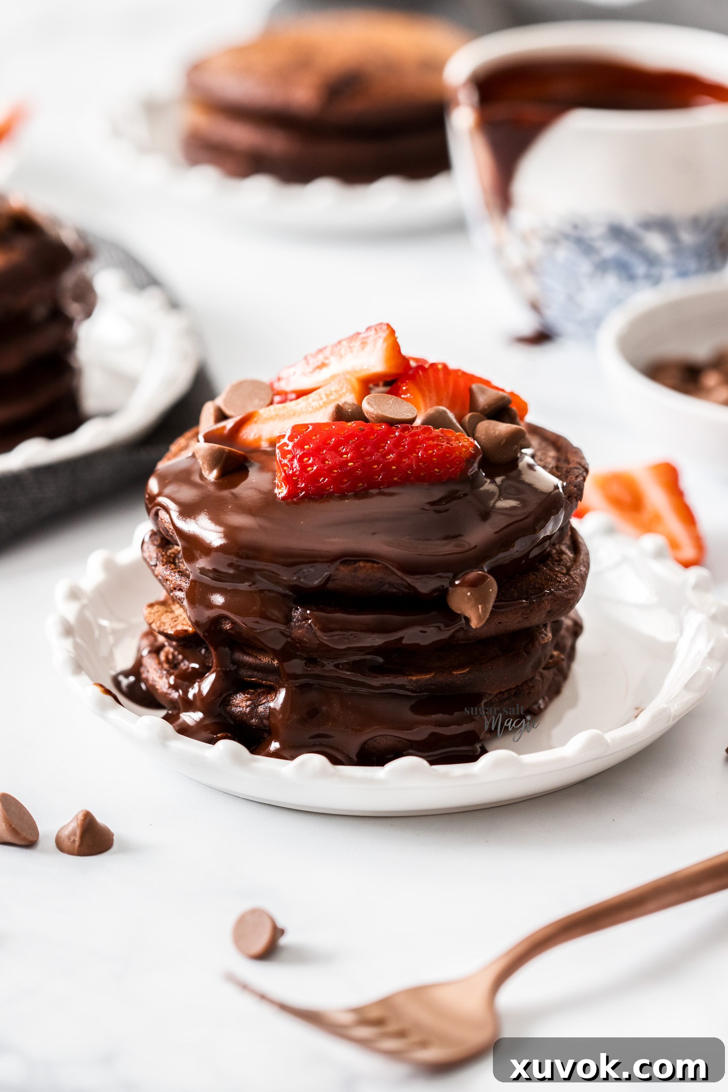A stack of 4 chocolate pancakes on a small plate, drizzled with chocolate sauce.