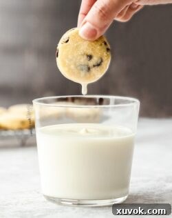 A glass of milk with a chocolate chip shortbread cookie being held above it and a drip of milk falling