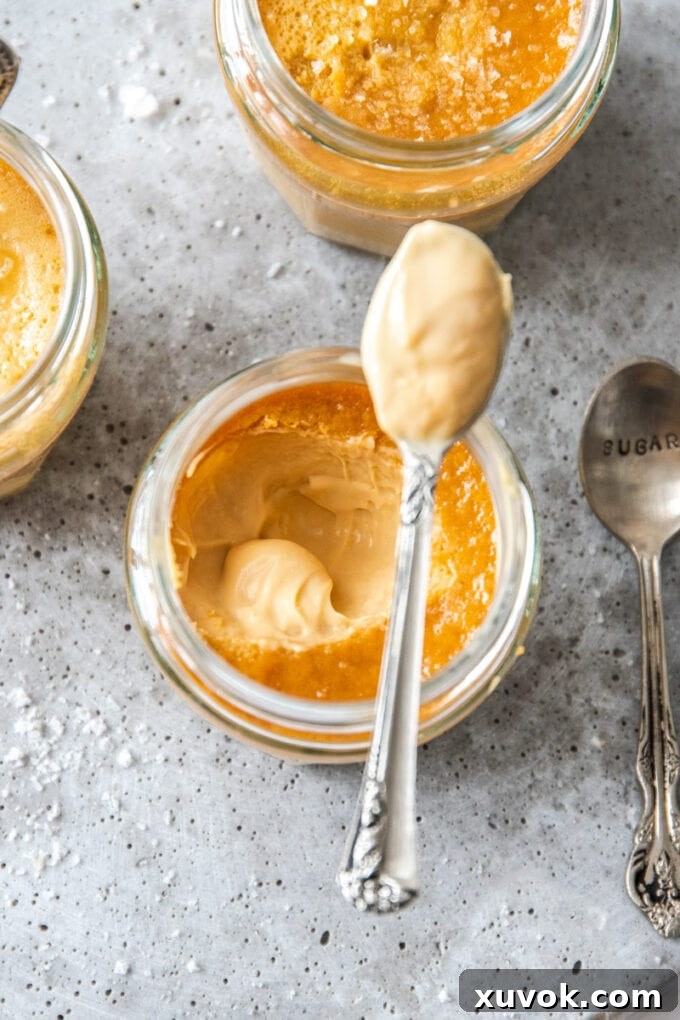 A small glass jar filled with luscious salted caramel custard, with a spoon resting elegantly on top, ready to be savored. This classic French dessert, Pots De Crème, offers a silky smooth texture and rich caramel flavor with a perfect touch of salt.