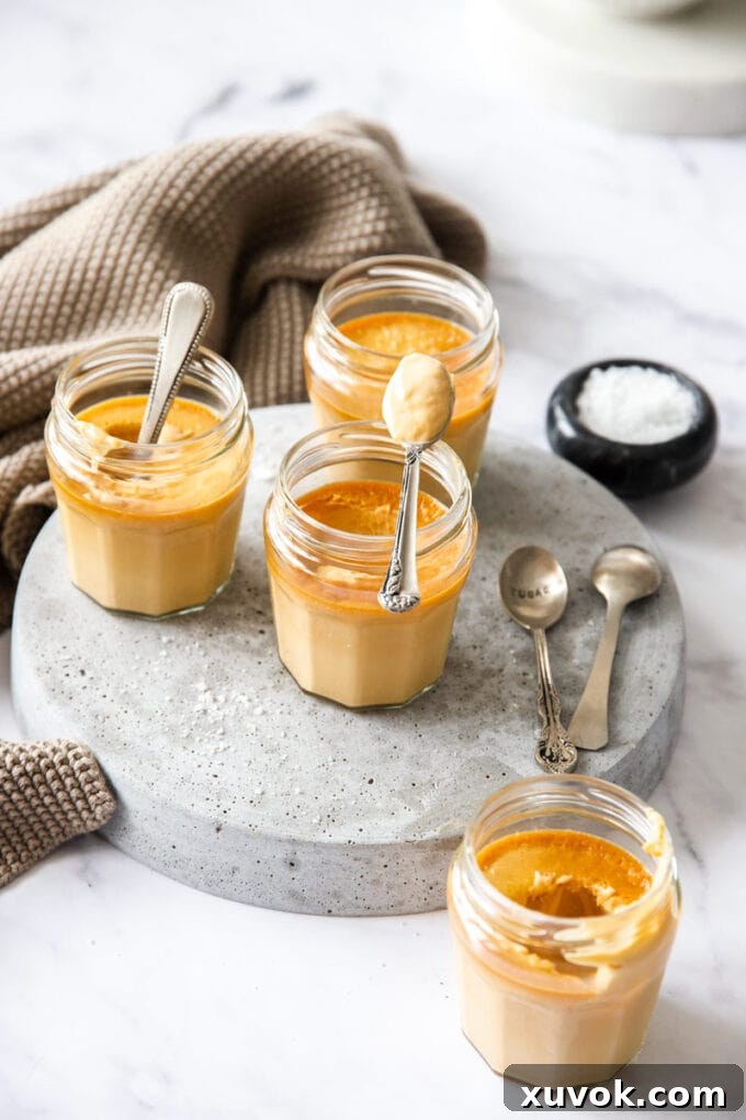 Several small glass jars filled with rich, glossy salted caramel custard, neatly arranged on a grey platter, ready for chilling or serving. The smooth surface of the custard promises an exquisite texture.