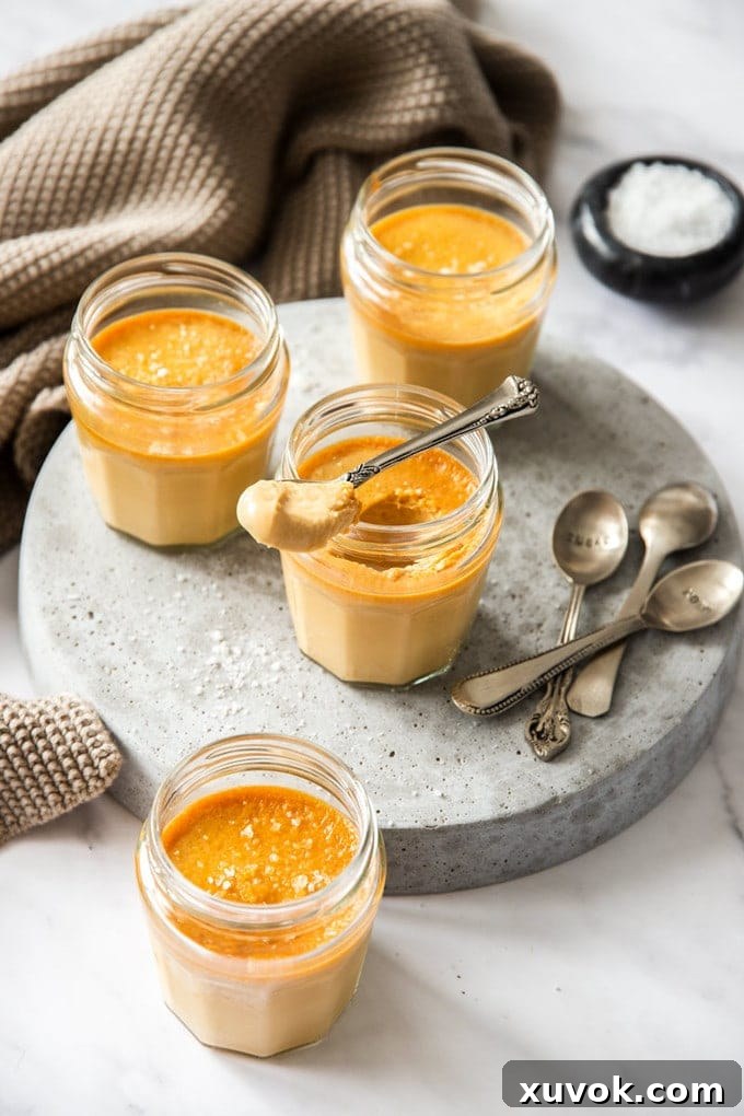 Small glass jars on a grey platter, filled with caramel custard. One has a spoon sitting on top, inviting a taste of the silky smooth French dessert.