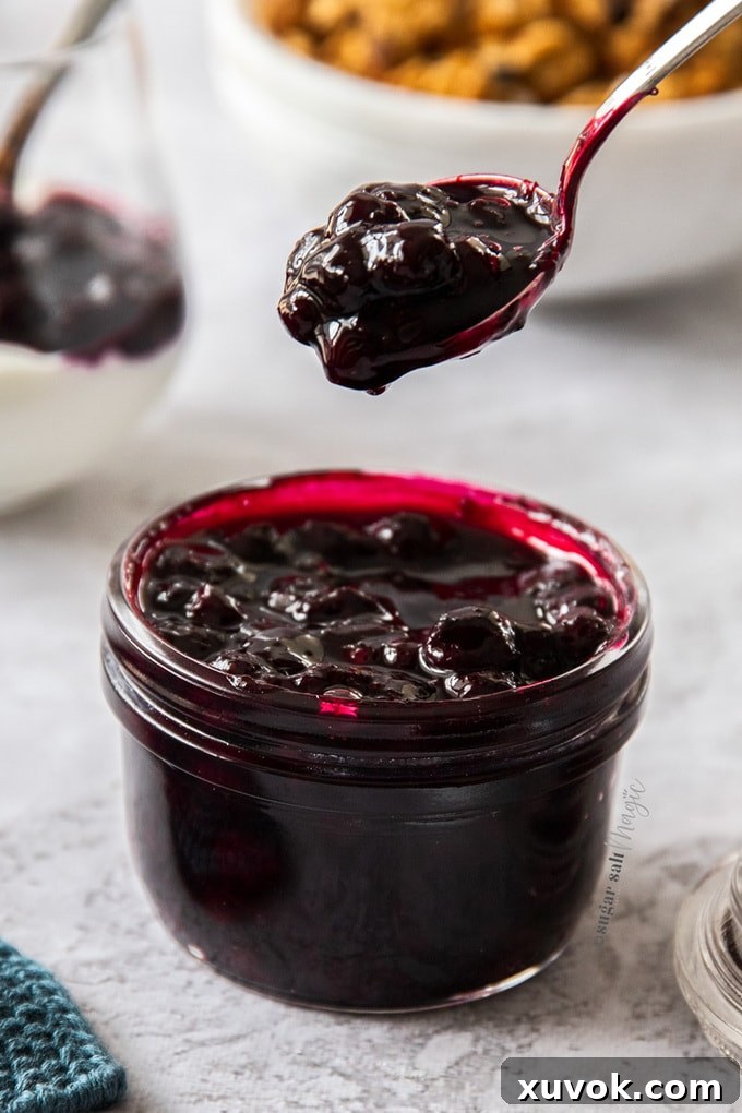 A small glass jar, filled to the brim with homemade blueberry pie filling. A spoon filled with it hovers above.