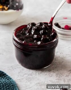 A small glass jar, filled to the brim with blueberry pie filling with a spoon digging in.