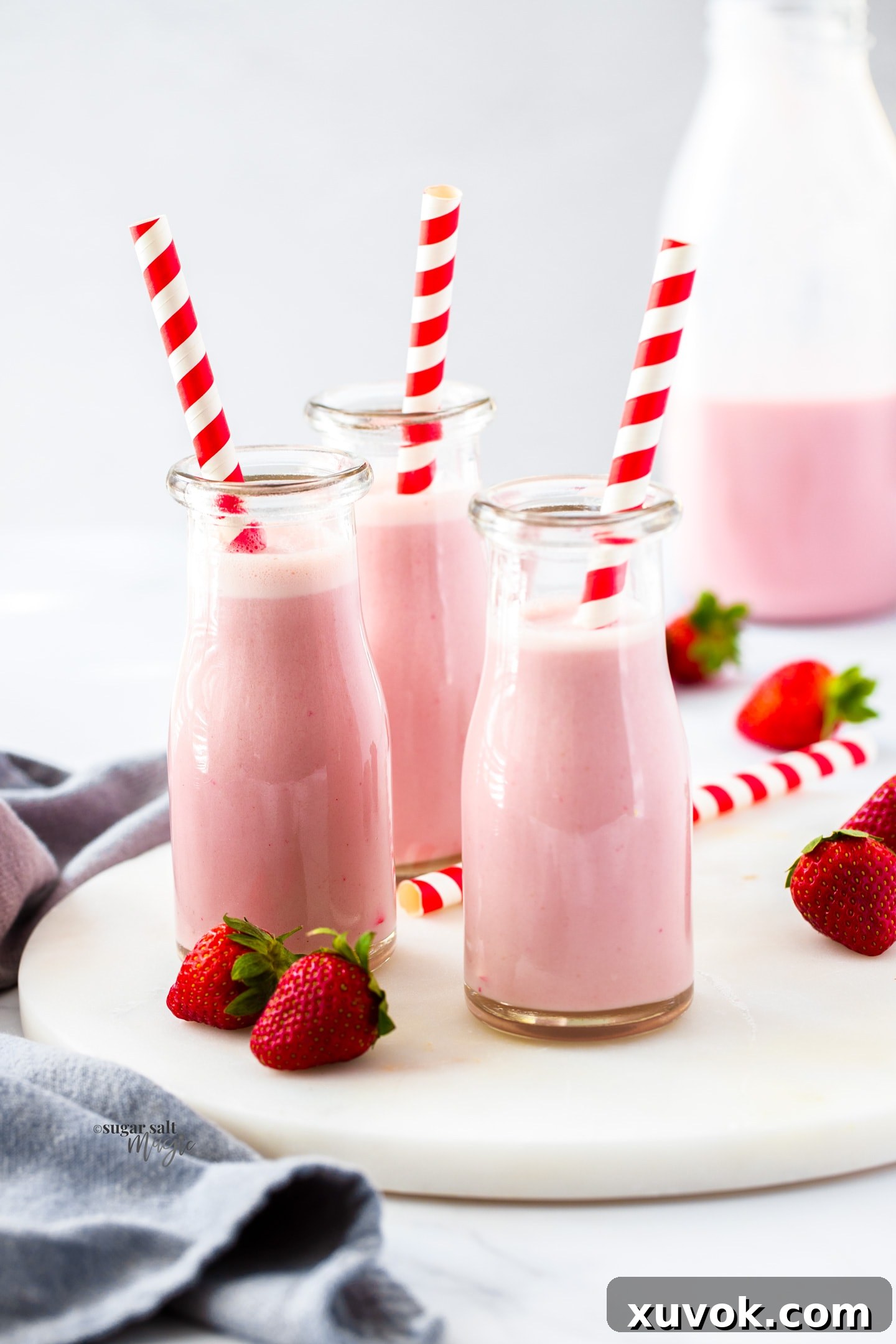 Three miniature milk bottles filled with vibrant strawberry milk, each adorned with a striped red straw, presented on a rustic wooden surface.