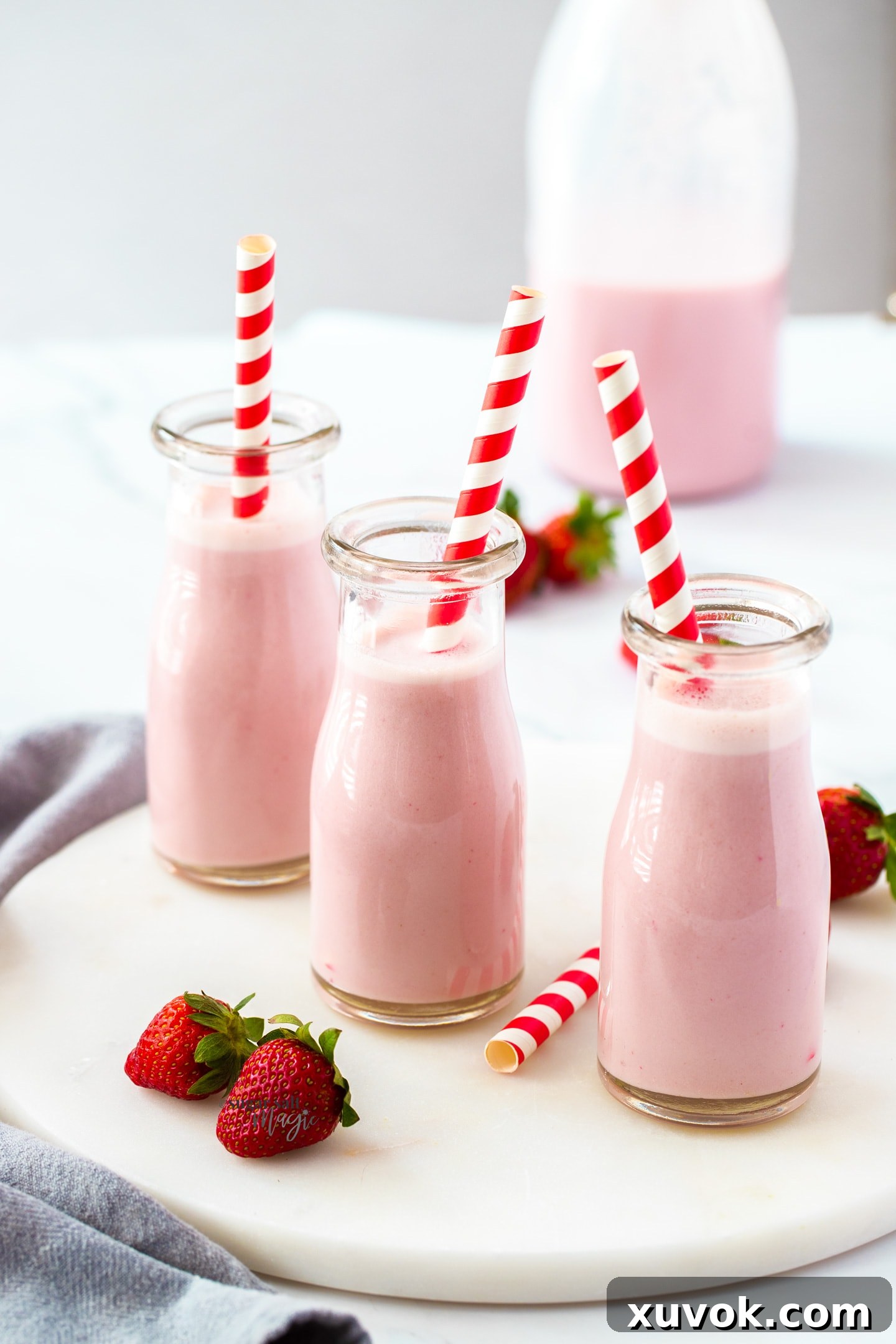 Three miniature milk bottles filled with vibrant strawberry milk, each with a striped red straw, arranged artfully with fresh strawberries.