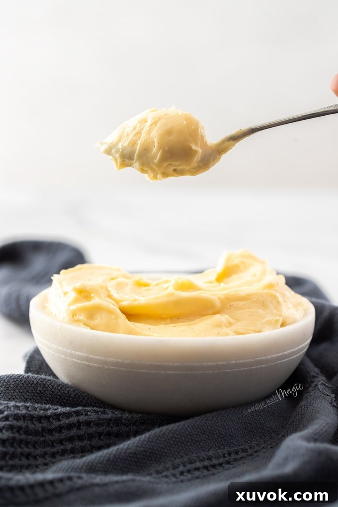 A pristine white bowl brimming with velvety vanilla pastry cream, with a spoon elegantly suspended above, holding a dollop of the rich custard.