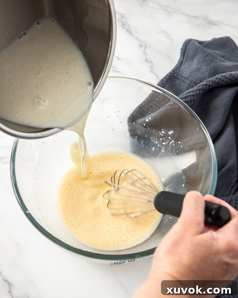 A steady stream of hot milk being carefully poured from a saucepan into a bowl containing the beaten egg and cornstarch mixture.