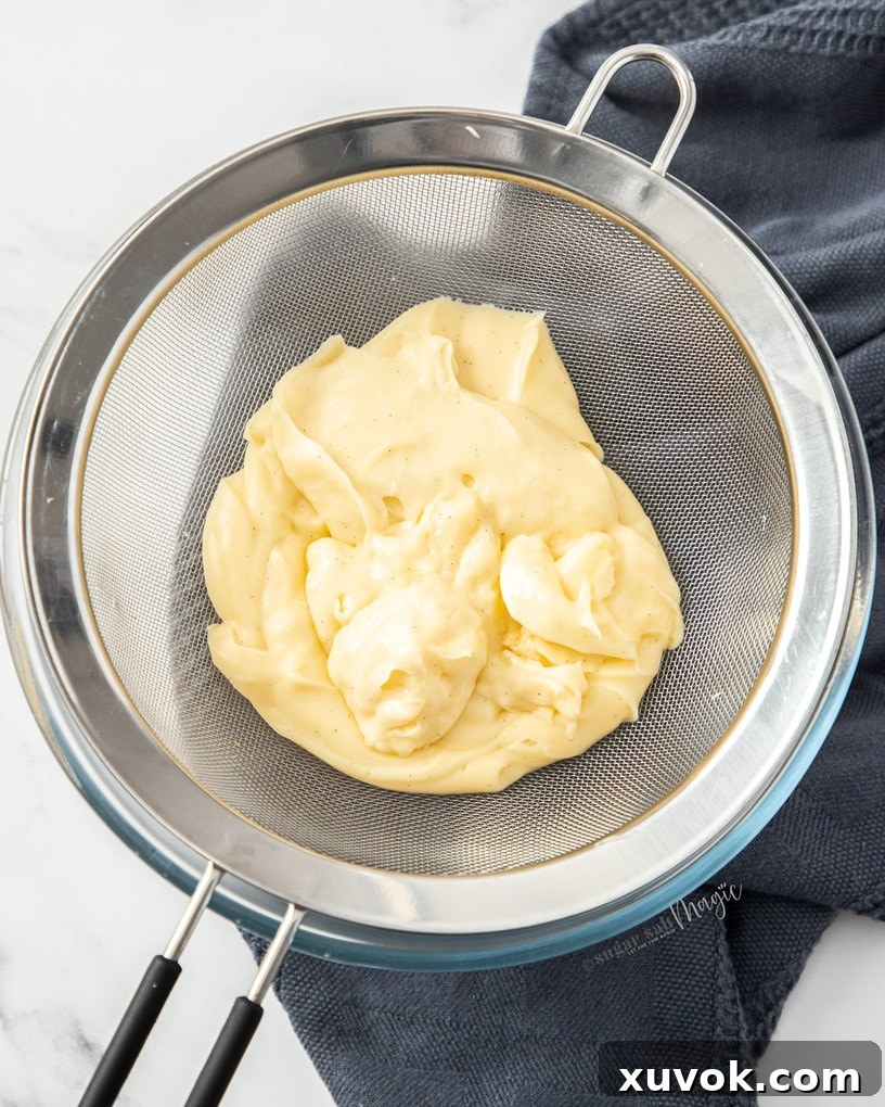 Rich vanilla pastry cream being strained through a fine-mesh sieve into a clean glass bowl, ensuring a perfectly smooth texture.