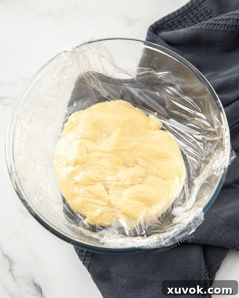 A glass bowl filled with freshly made pastry cream, tightly covered with plastic wrap pressed directly onto its surface to prevent a skin from forming.