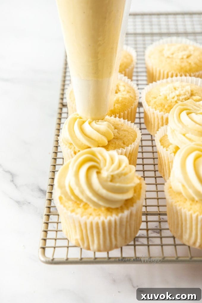 A row of vanilla cupcakes on a wire rack with smooth German buttercream being piped on top, showing elegant swirls.
