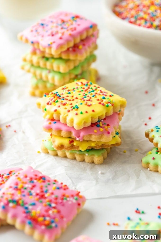 Stacks of brightly colored iced sugar cookies covered in sprinkles, arranged on a sheet of baking paper.