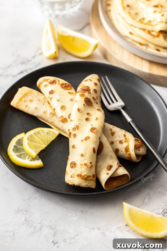 English pancakes rolled and stacked on a black plate with lemon wedges nearby.