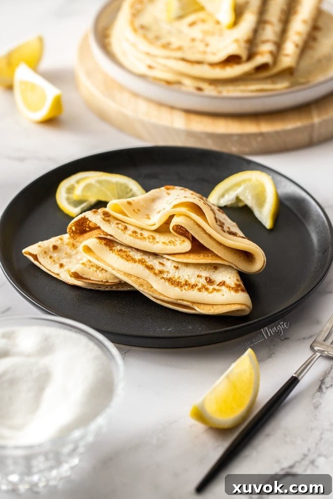 English pancakes folded and stacked on a black plate with lemon wedges nearby.