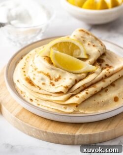 A white plate topped with a stack of english pancakes and wedges of lemon
