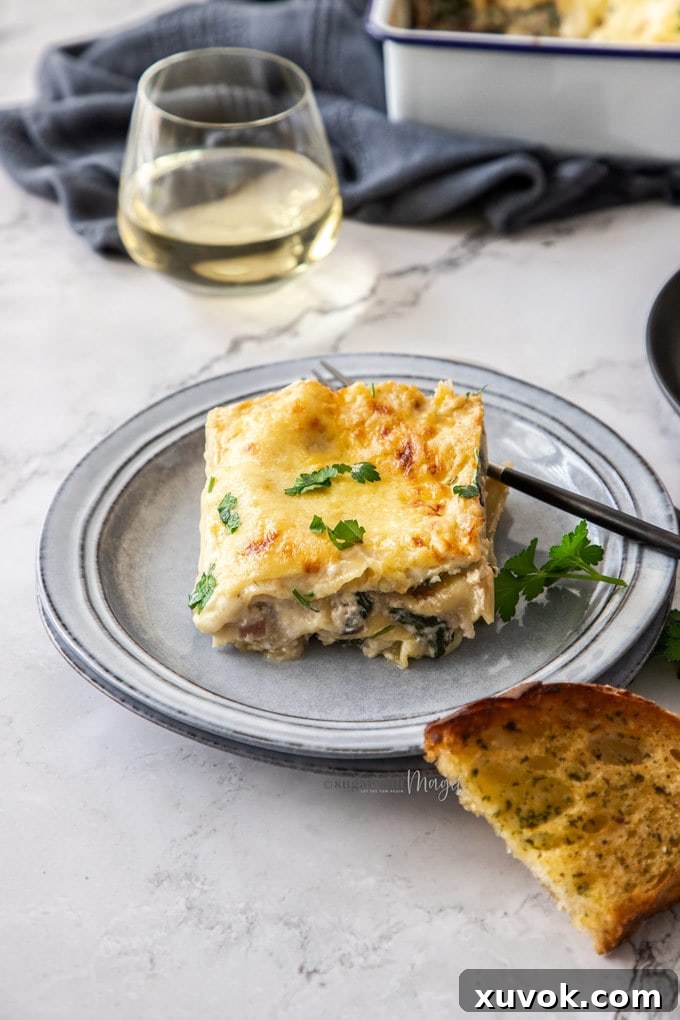 A perfectly portioned slice of white chicken lasagna, showcasing its creamy layers of pasta, chicken, spinach, and mushrooms, served on a grey plate with garlic bread in the foreground and a glass of wine in the background.