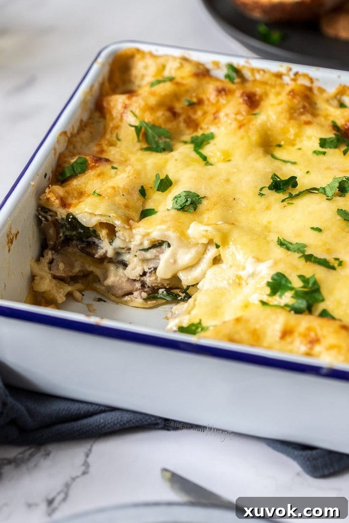 A close-up view of a slice of white chicken lasagna, revealing its perfectly cooked layers of pasta, chicken, spinach, mushrooms, and creamy cheese sauce, nestled in a white baking dish.