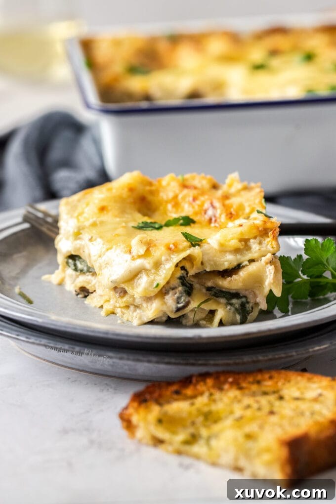 A glistening slice of white chicken lasagna, impeccably layered with creamy sauce, chicken, spinach, and mushrooms, served on a grey plate alongside a piece of golden garlic bread.