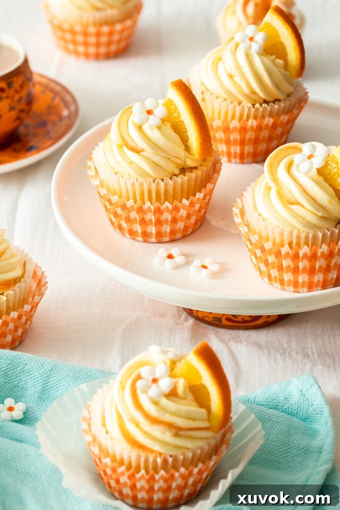 Three orange cupcakes with white buttercream on a white plate, garnished with orange wedges and tiny fondant blossoms.