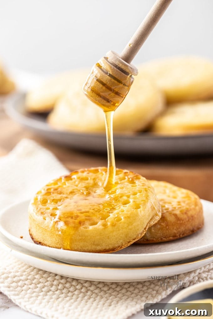 Two golden homemade crumpets on a white plate with rich honey being drizzled over them, highlighting their iconic porous texture.