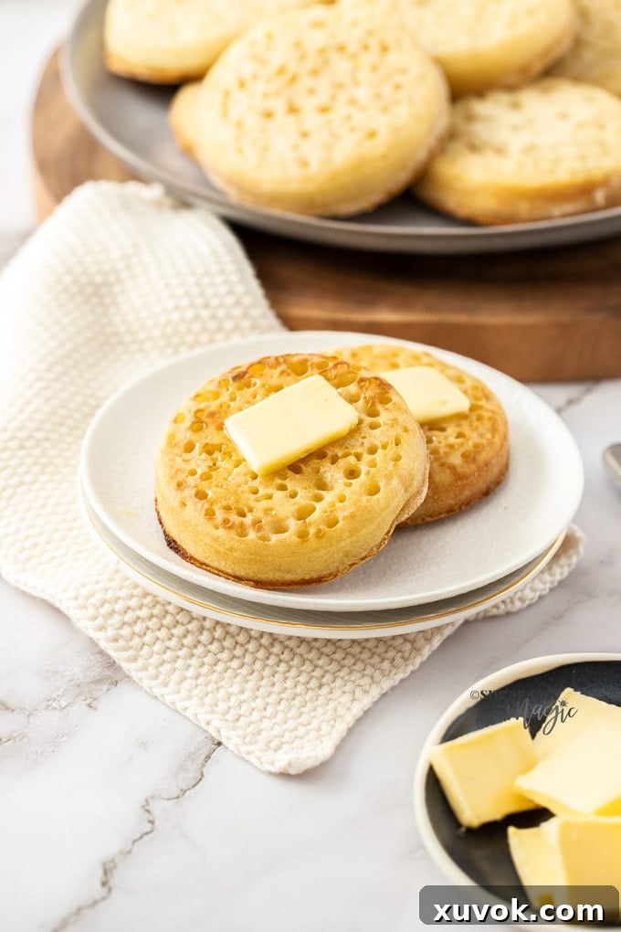 Two golden-brown homemade crumpets on a pristine white plate, generously spread with melting butter, ready to be enjoyed.