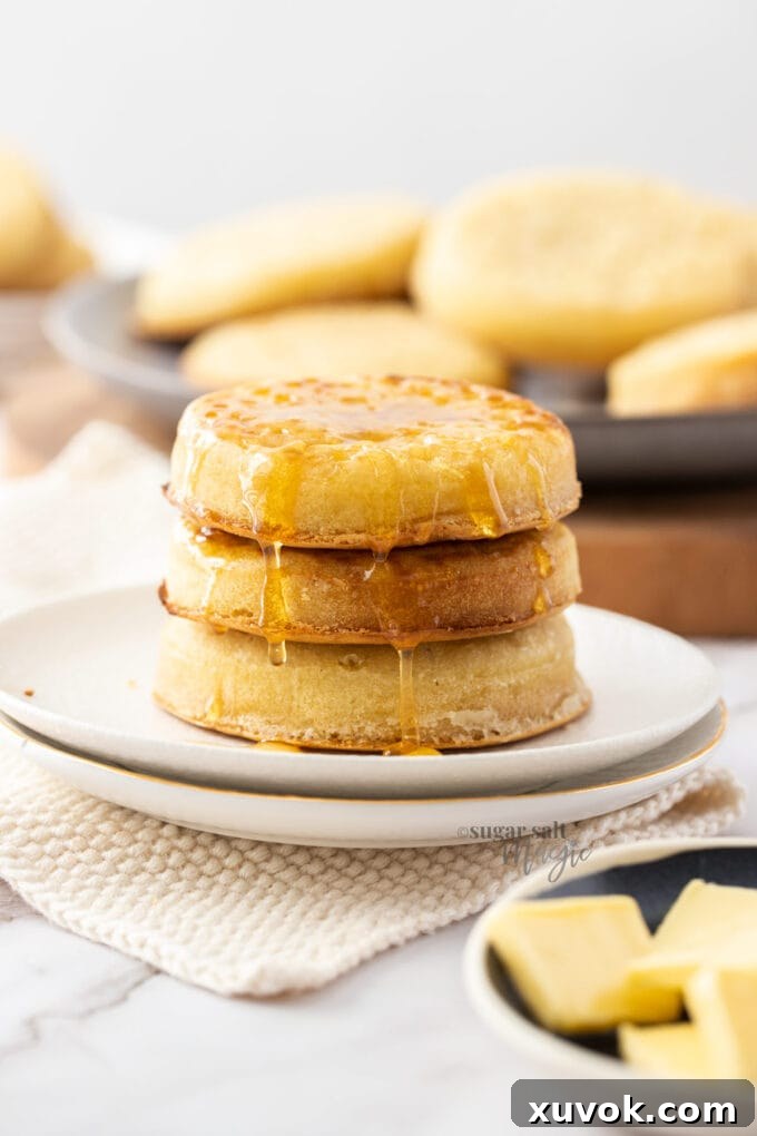 Three perfectly stacked homemade crumpets on a white plate, with more crumpets visible in the soft background.
