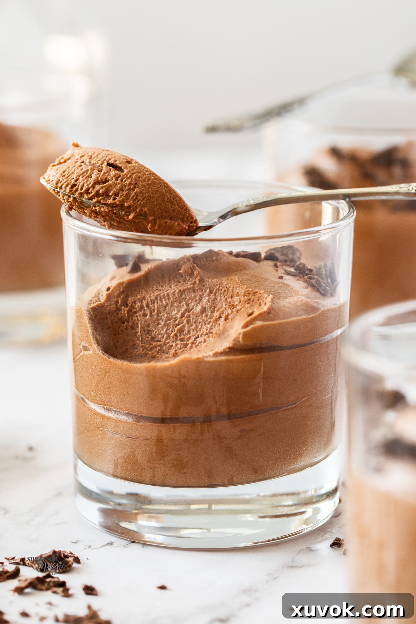 A glass of chocolate mousse with a spoon full of chocolate mousse laying across the top of the glass, garnished with chocolate shavings.