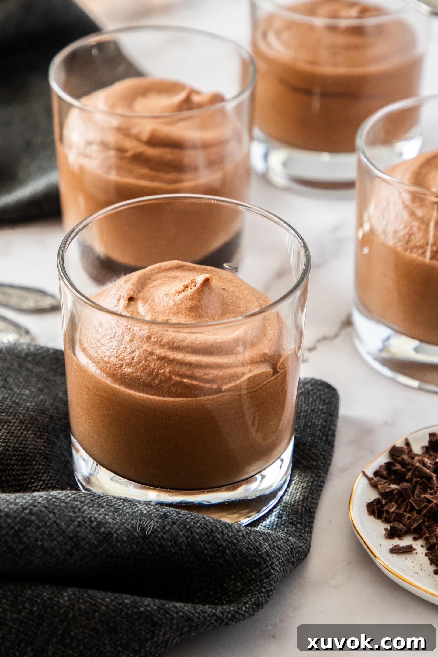 Four glasses of eggless chocolate mousse with dark serviettes, a plate of chocolate shavings, and a sprig of mint next to them.