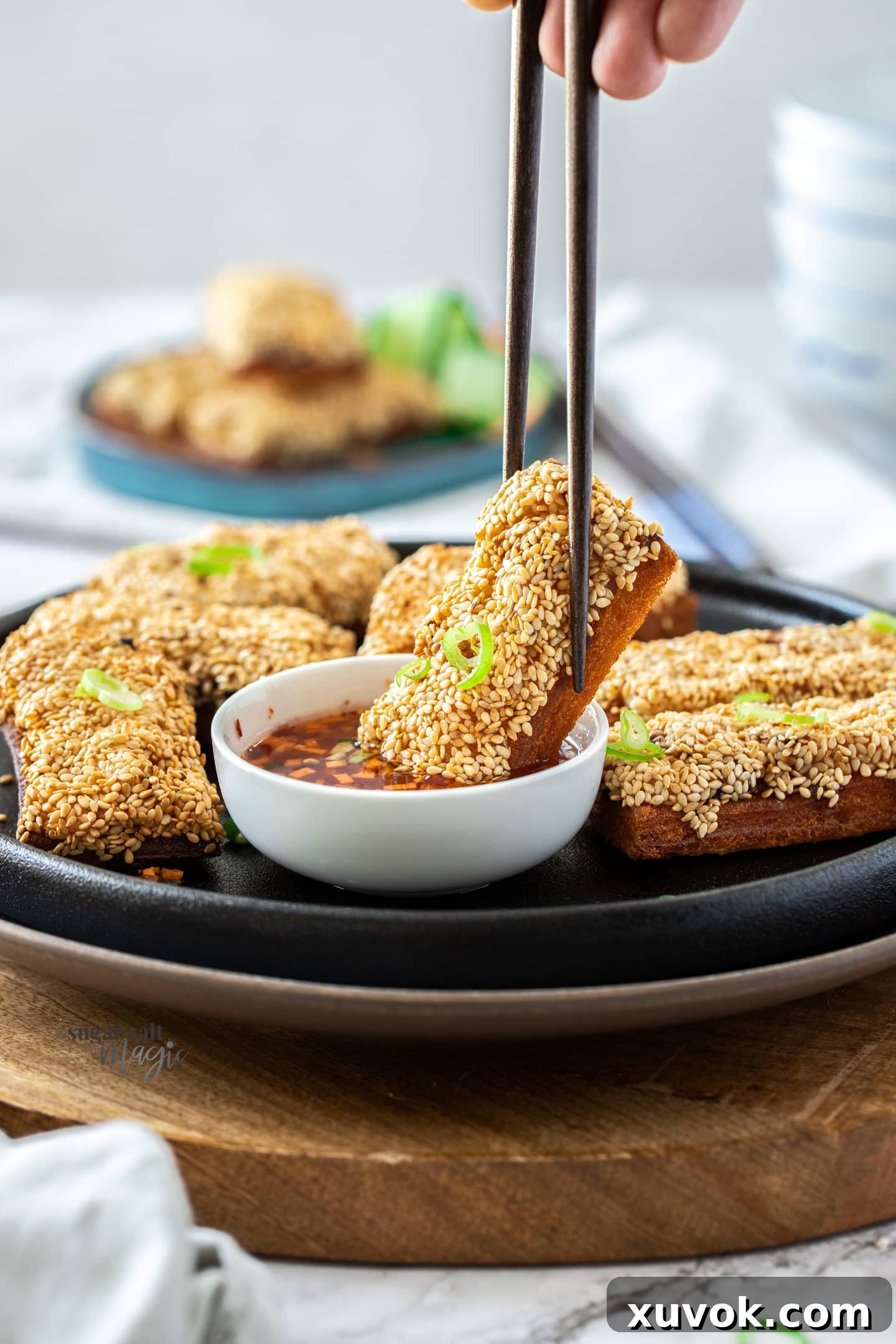 A slice of crispy sesame prawn toast being dipped into a small white bowl of sweet chili sauce, highlighting its golden texture.