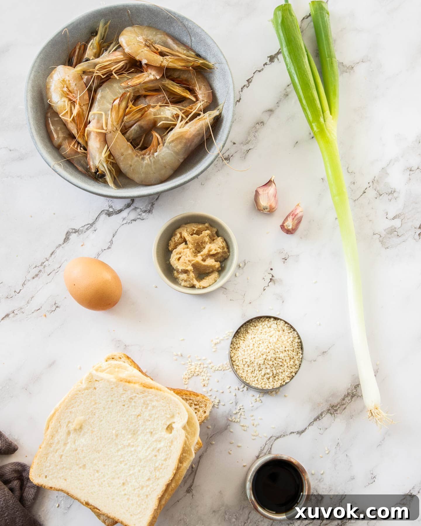 Fresh ingredients for homemade prawn toast laid out on a marble surface, including raw prawns, bread slices, and sesame seeds.