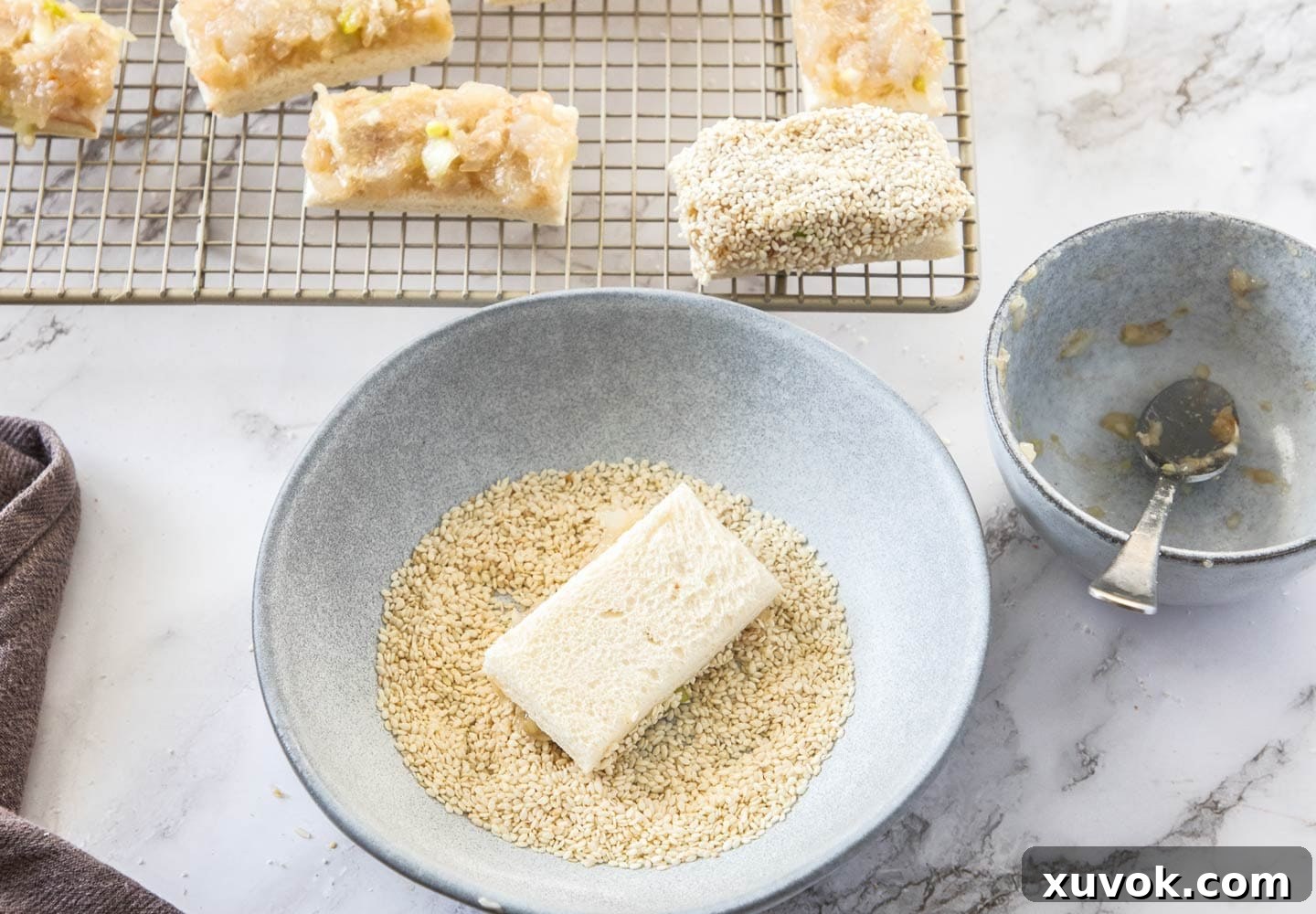 A piece of prawn toast being dipped, prawn-side down, into a bowl of white sesame seeds to coat.
