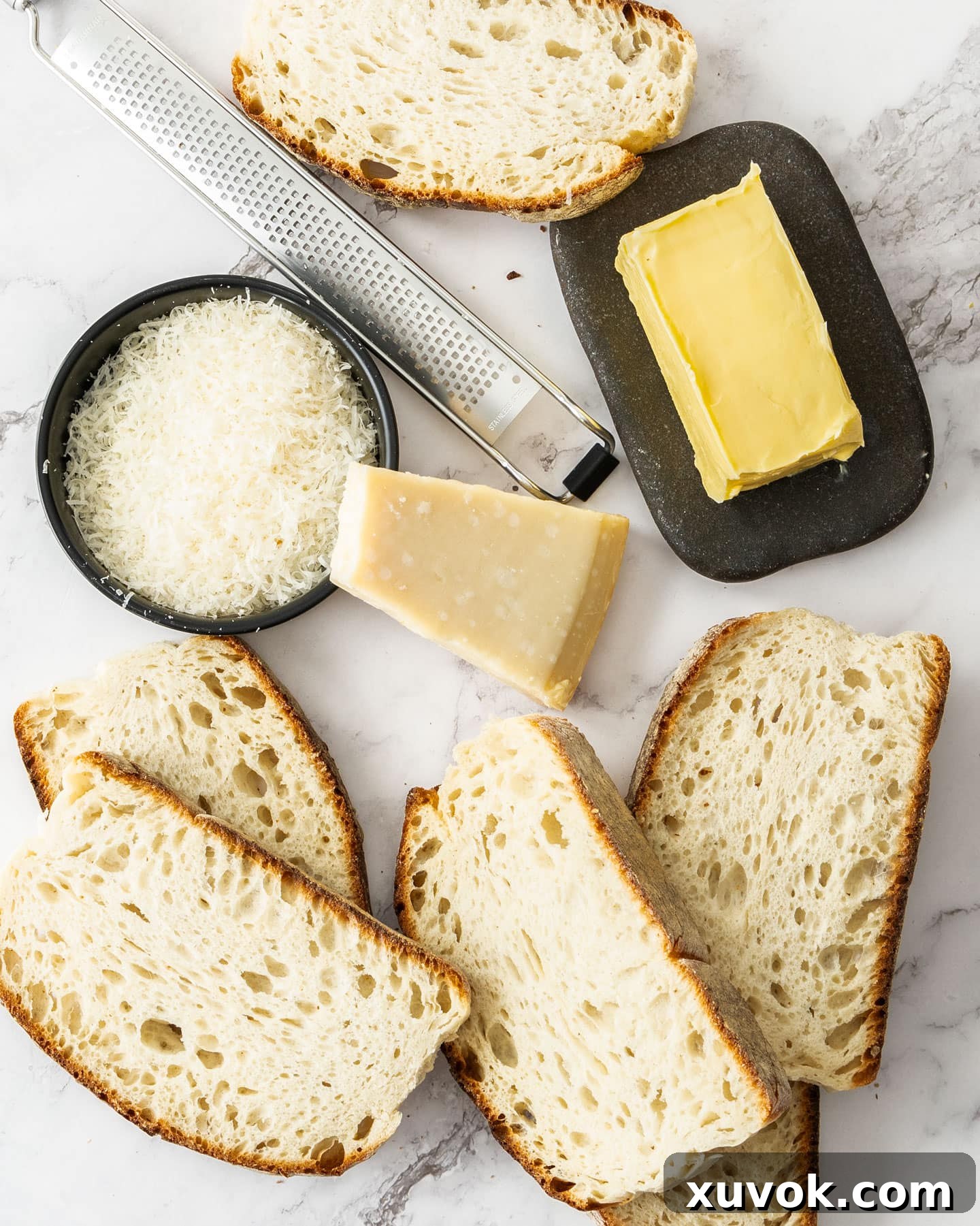 Ingredients for parmesan toast on a marble surface, including bread, butter, and a block of parmesan cheese.