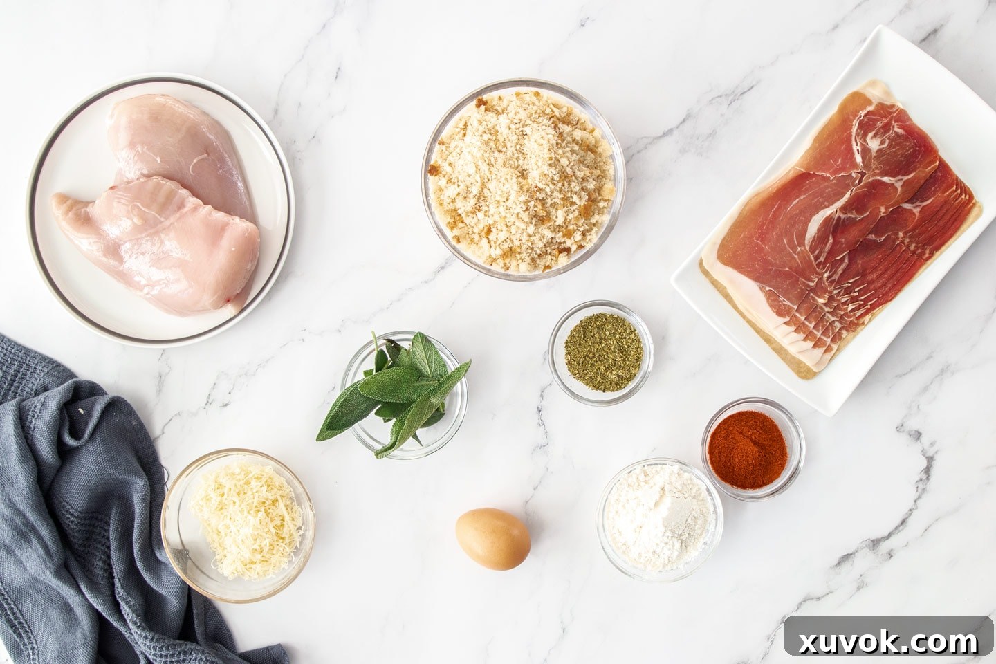 A flat lay of ingredients for Saltimbocca chicken schnitzel, including chicken breasts, fresh sage, prosciutto, parmesan, breadcrumbs, flour, and eggs.