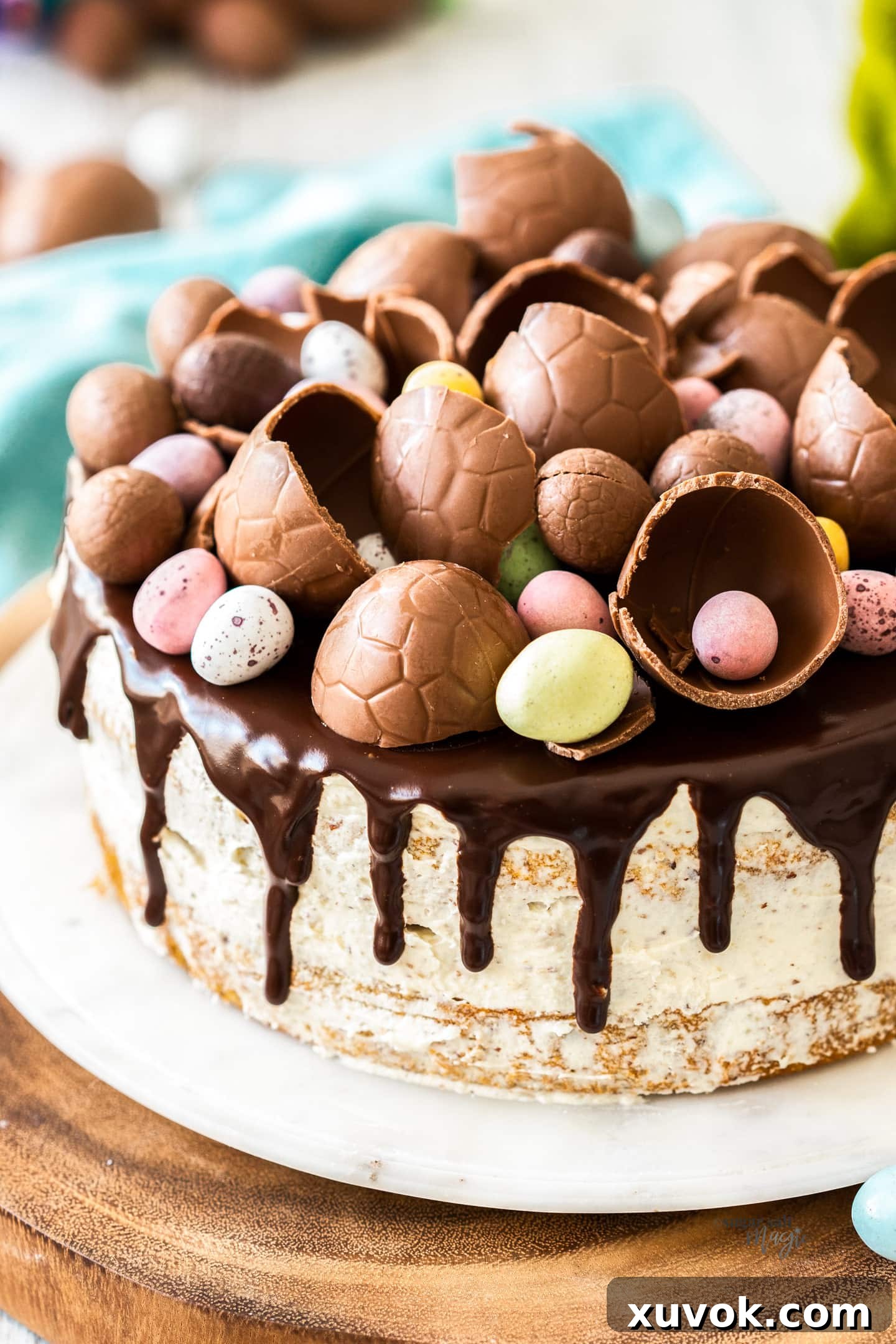 A beautifully decorated hazelnut cake on a cake platter, generously adorned with a glossy ganache and an abundance of colorful Easter eggs.
