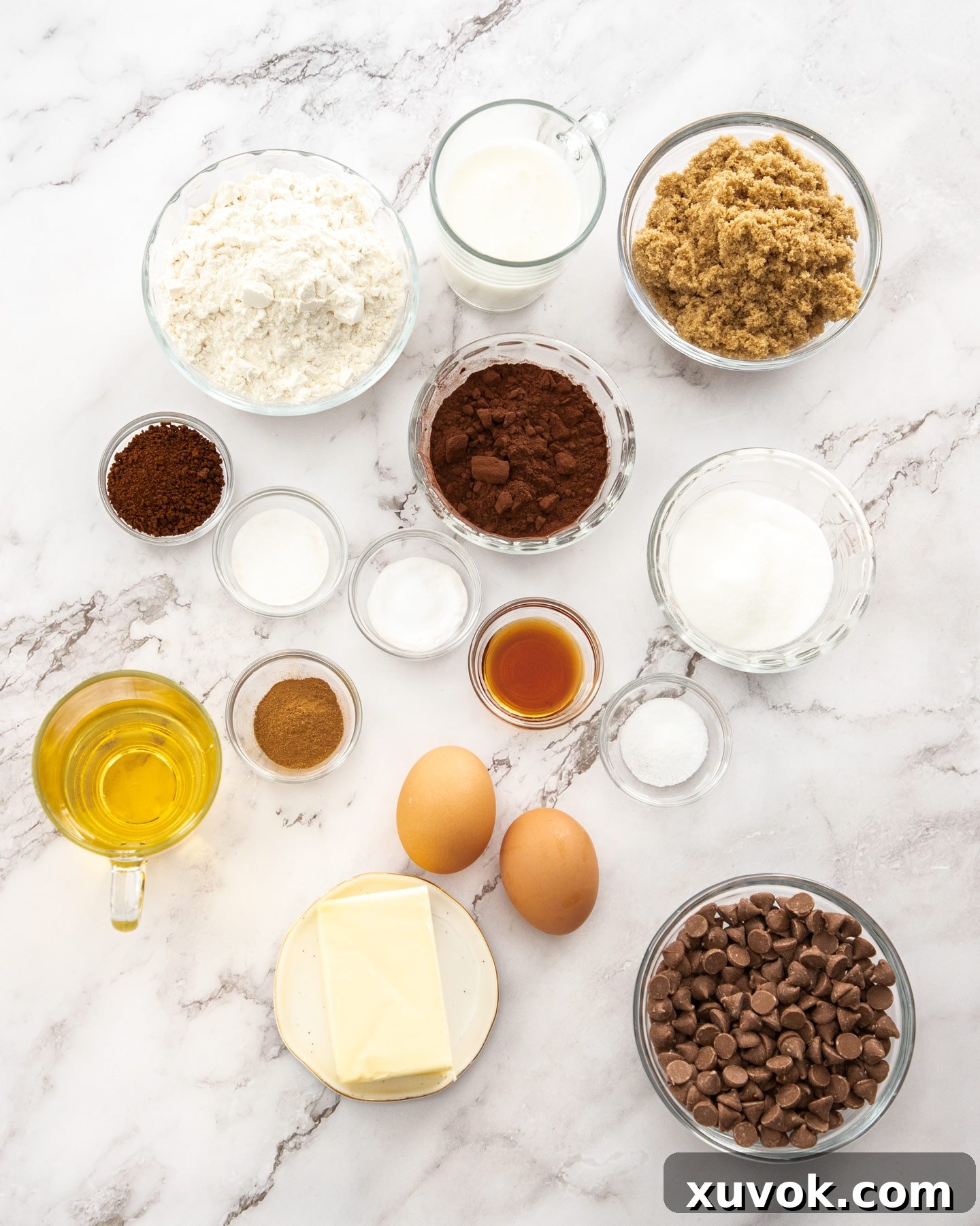 Ingredients for chocolate crumb cake, including flour, cocoa powder, sugars, butter, and wet ingredients, neatly arranged on a marble bench top, ready for baking.