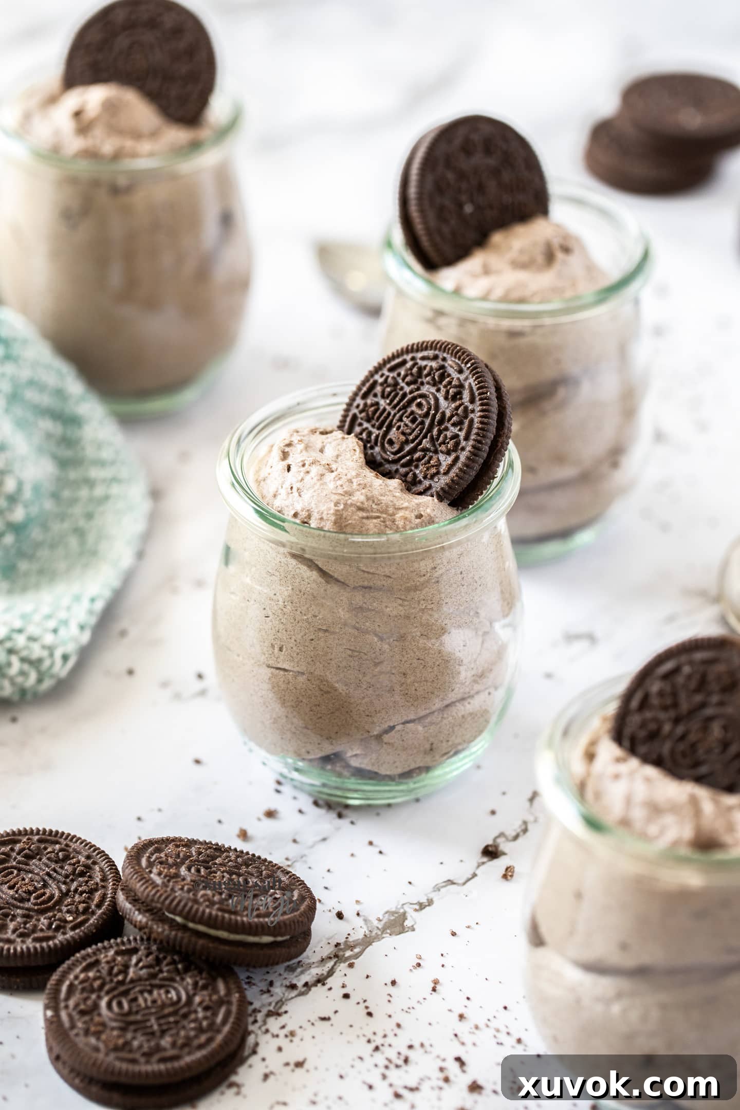Four elegant glass jars, each generously filled with creamy Oreo mousse, are artfully arranged on a light-colored surface. A single, perfectly placed Oreo cookie crowns each dessert, hinting at the delicious cookies and cream flavor within. The setting is bright and inviting, showcasing the dessert's delightful texture.