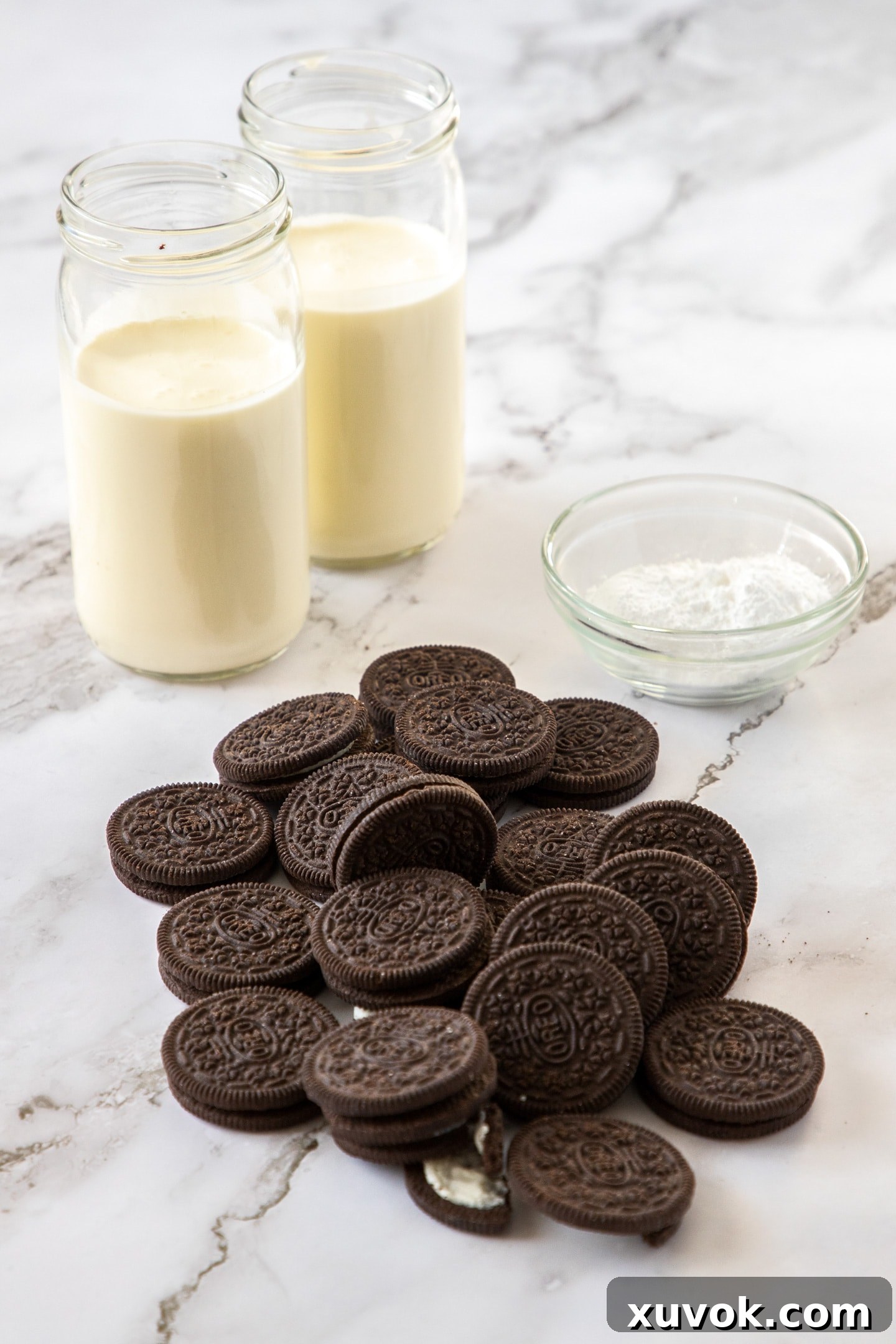 The three core ingredients for this simple Oreo mousse: a stack of classic Oreo cookies, a clear bowl filled with fresh heavy cream, and a small white dish holding fine icing sugar. They are neatly arranged on a pristine white marble countertop, suggesting a clean and straightforward recipe.
