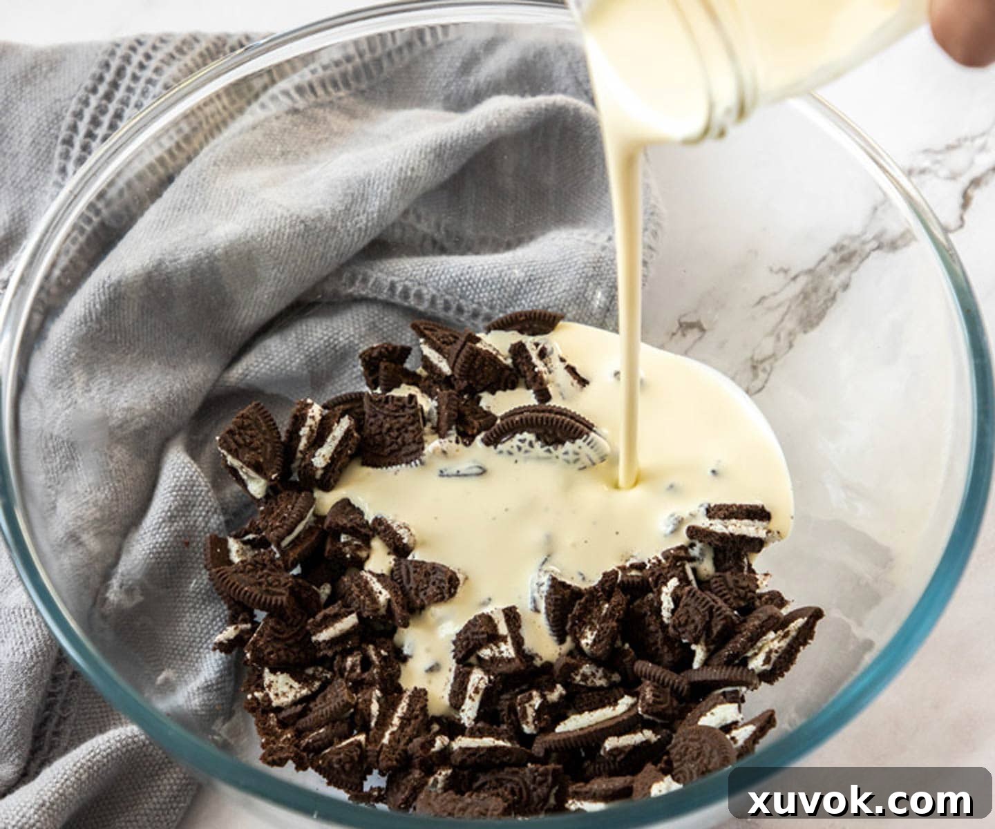A large glass bowl containing roughly chopped Oreo cookies, with rich, white heavy cream being poured over them. The scene suggests the beginning of the overnight soaking process, essential for this no-bake Oreo mousse.