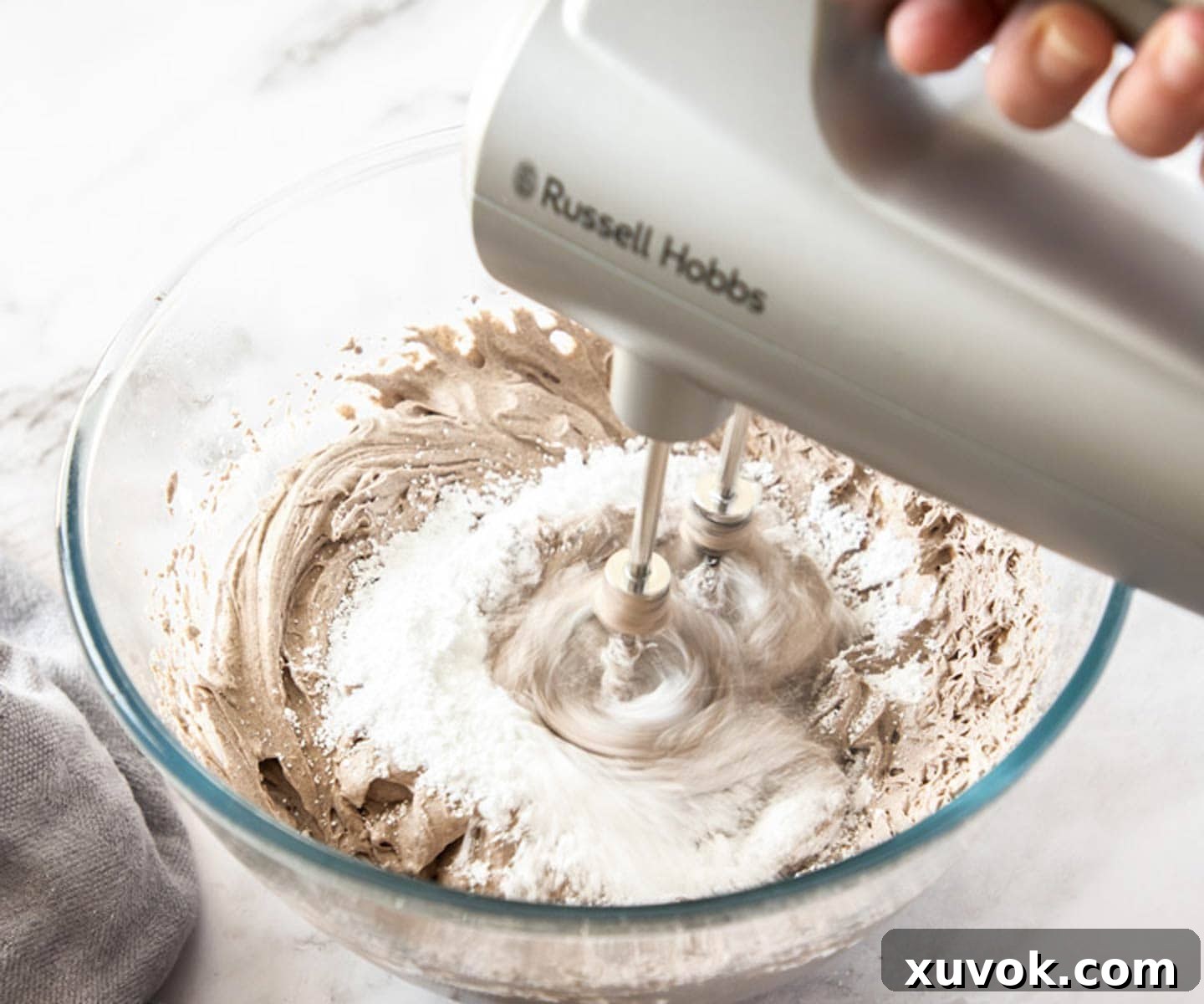 A hand is shown whisking fine powdered sugar into a bowl of already whipped Oreo cream mixture. The mousse appears smooth and speckled with cookie pieces, ready for final sweetening.
