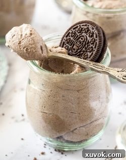 A close-up shot of a single glass jar brimming with fluffy 3-Ingredient Oreo Mousse, adorned with a whole Oreo cookie on top and a spoon resting casually on the rim, inviting a taste. The focus is sharp, highlighting the creamy texture and cookie specks.