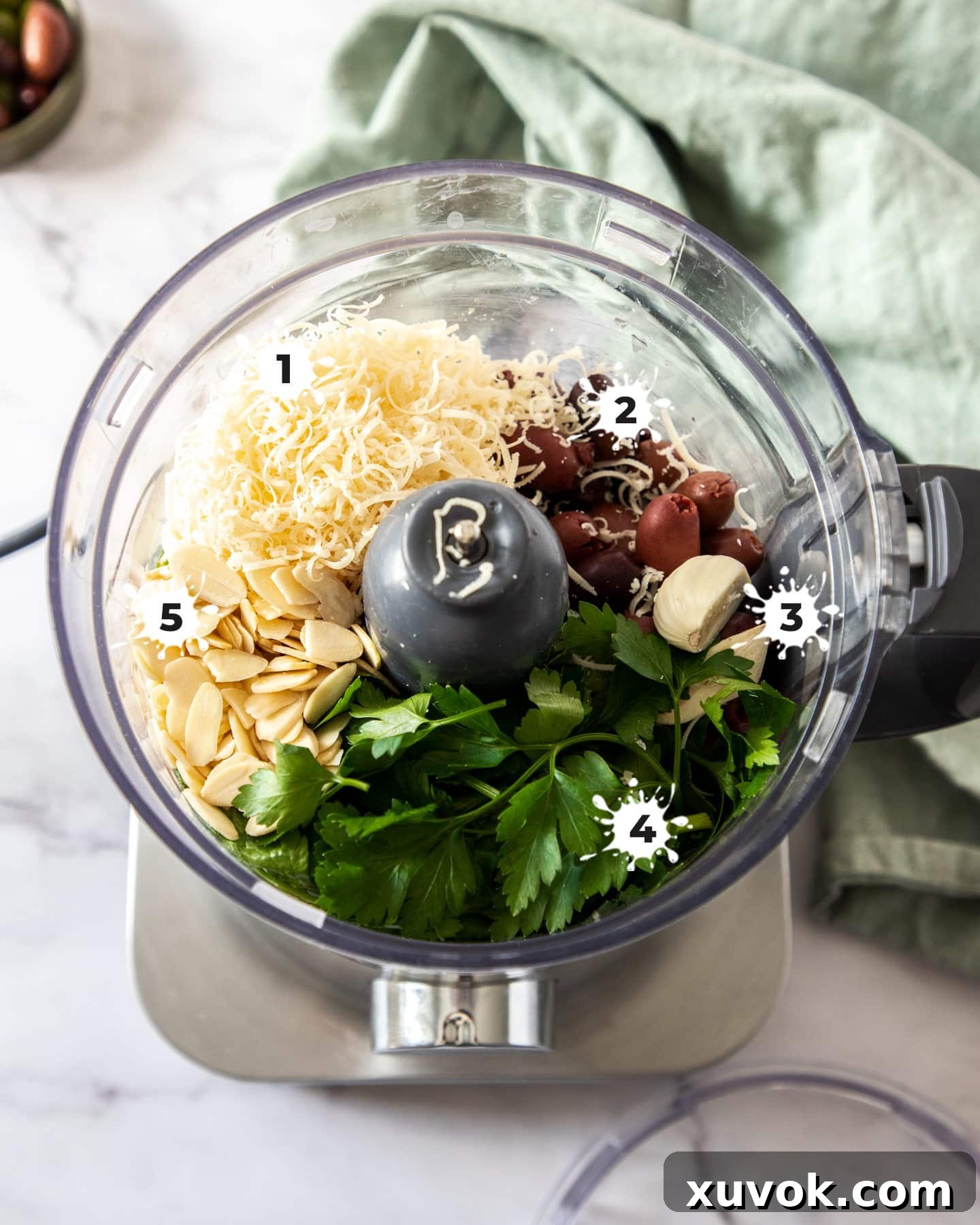 A food processor bowl filled with fresh parsley, pitted Kalamata olives, garlic, parmesan cheese, and almonds, ready to be transformed into homemade olive pesto.