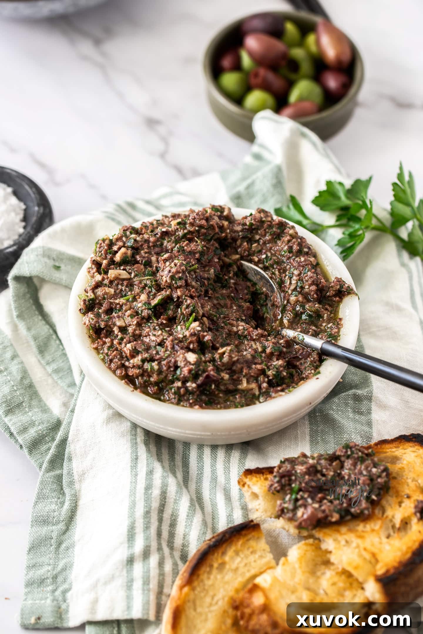 A pristine white bowl filled with creamy Kalamata olive pesto, with a side of fresh olives and toasted bread, inviting a delightful culinary experience.