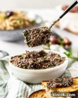 A spoon with rich olive pesto hovering over a white bowl, with crusty bread nearby, ready to be served.