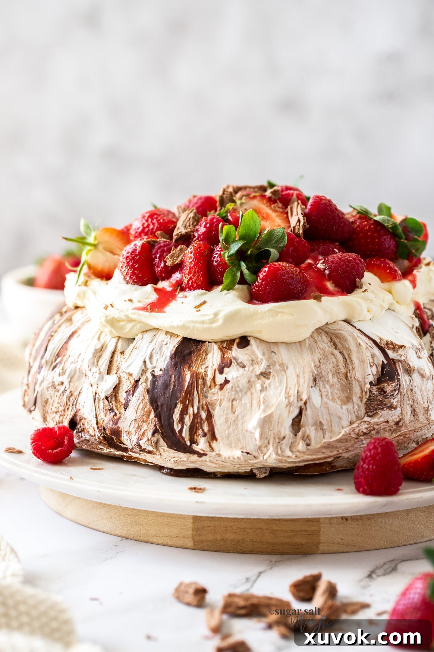 A chocolate pavlova topped with cream and berries on a white cake plate