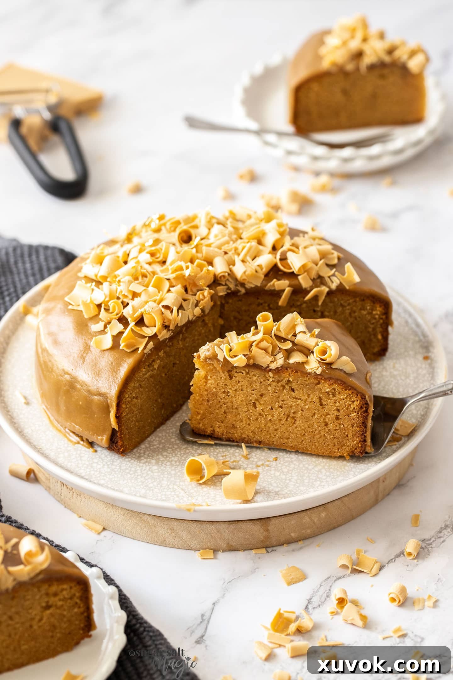 A beautifully decorated caramel mud cake topped with white chocolate shavings, presented on a pristine white plate, showcasing its golden-brown hue and enticing texture. Perfect for any celebration.